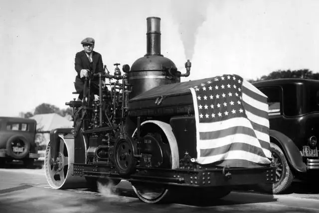 In 1929, the opening of this paved road was grand enough to warrant a celebration, as shown in the photograph. This patriotically decorated machine is a true steamroller (powered by a steam engine, at center)âand operated with Queens class by a Queens public works commissioner J. J. Halloran, who wears a three-piece suit and a tie.